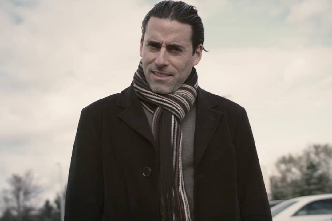 A man in a black coat, and a scarf stood in front of a car